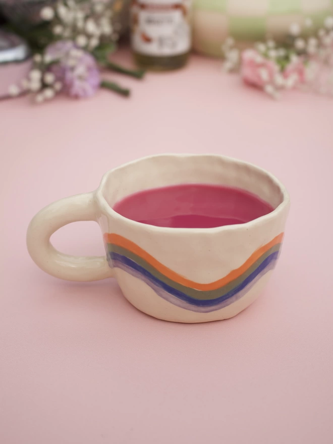 wavey stripes of orange, green, blue and purple around a handmade stoneware pottery mug with bright pink liquid inside