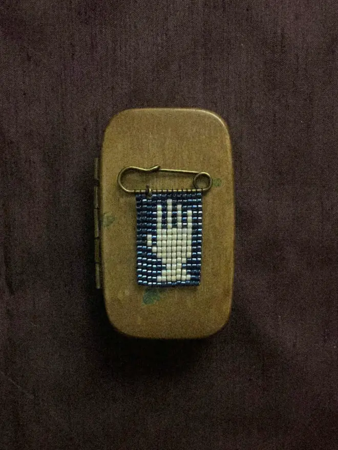 A beaded pin badge with a white hand on a blue background. The pin is sitting on a brass box.