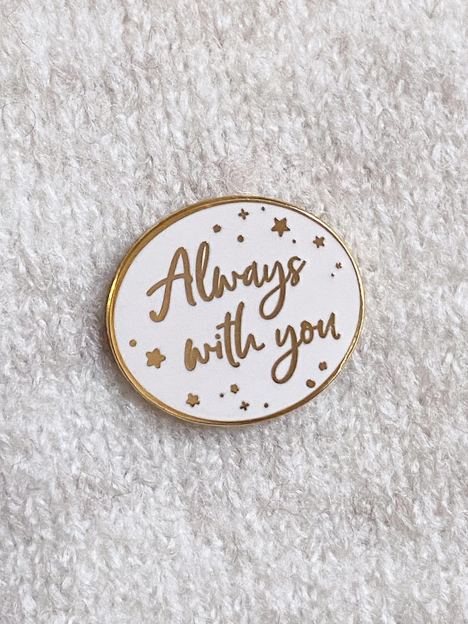 A white and gold enamel pin badge is pinned to a beige jumper. It has a subtle star design and the words "Always with you".