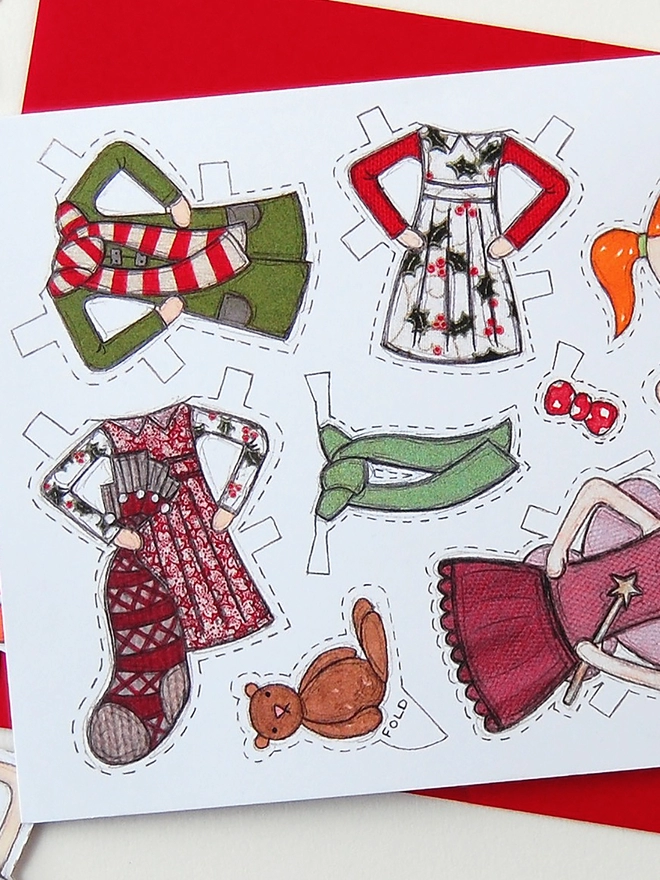 A Christmas card with an illustrated paper doll design is with a red envelope on a white desk.