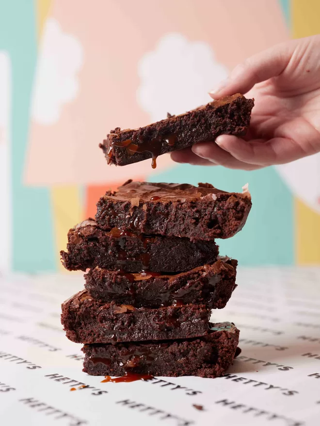 Sea salted caramel brownies in a stack with a hand adding another brownie to the top of the pile with caramel dripping in front of a colourful background