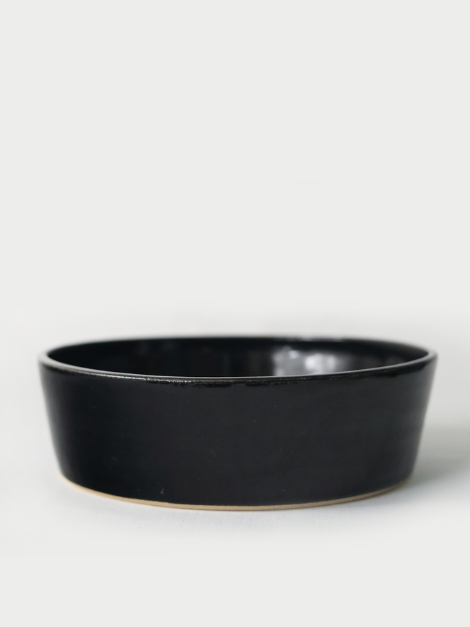 A single wide, shallow glazed ceramic pasta bowl in jet black, photographed from a slight angle against a plain light grey background, showing the unglazed natural clay base.
