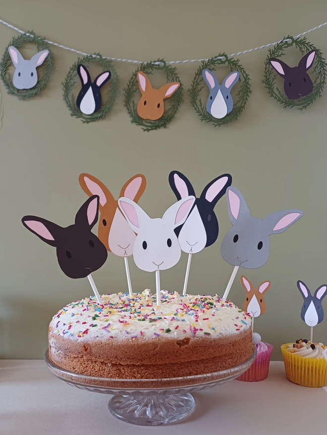 12 Bunny Easter Garland displayed on wall behind large cake featuring 5 Easter Bunny Cake Topper Decorations. Bunnies are a mix of grey, brown, black and white.