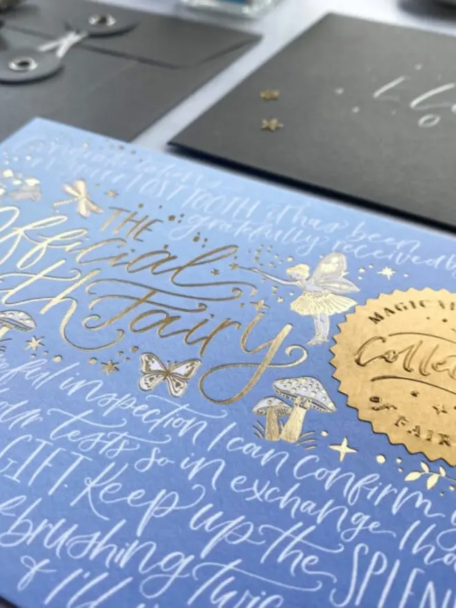 Close-up of the blue ombre notecard with gold foil "The Official Tooth Fairy" lettering and a fairy with a wand. Grey envelopes and an embossed gold seal are just in view.