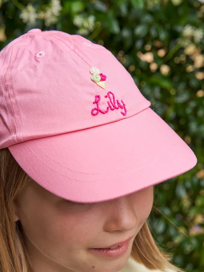 Hand embroidered personalised children's cap in pink. The pink cap is worn by a girl and the cap is embroidered with flowers. 