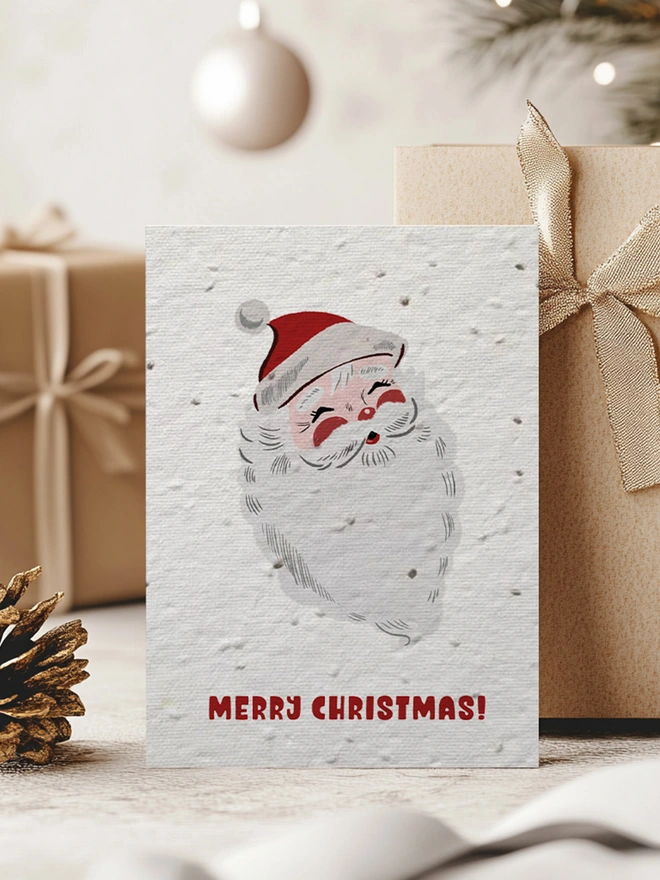 Christmas card with an illustrated Santa Clause with 'Merry Christmas' written underneath. In the background is presents in a Christmas scene