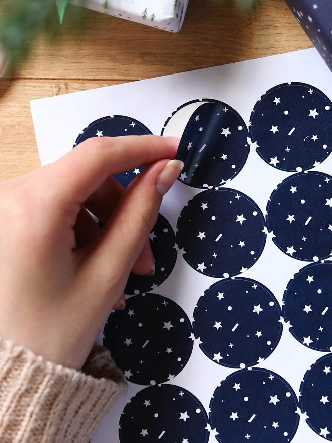 A hand is peeling a navy blue sticker with a tiny star pattern from a sheet of 35 matching stickers.