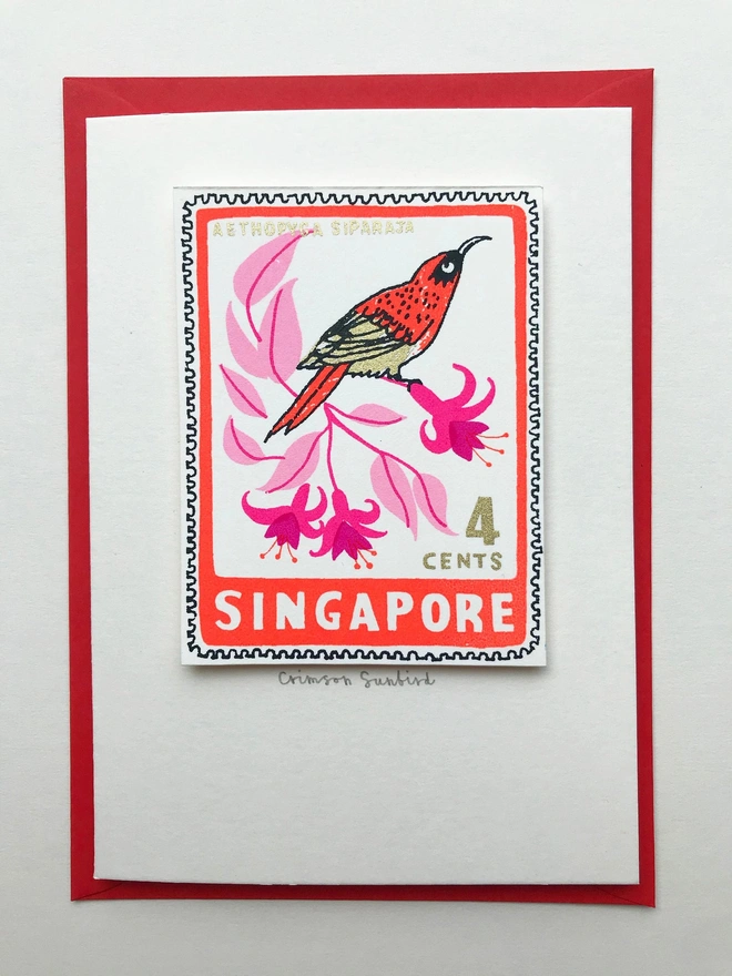 Bird Stamp Card Singapore/ Crimson Sunbird, a white card with a colourful bird design in the centre above a red envelope on top of a white surface. 