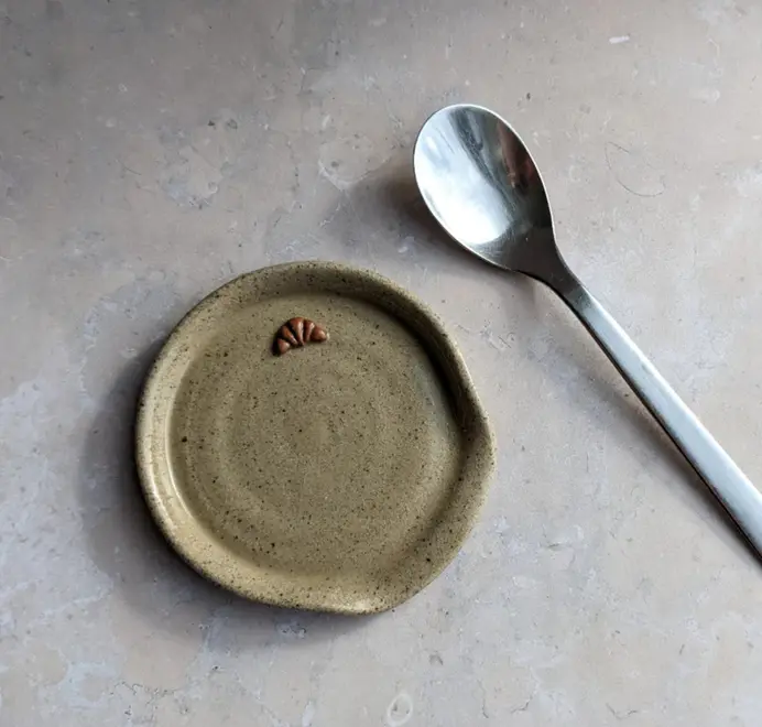 Stoneware spoon rest in biscuit glaze with a croissant design and a spoon beside it, on a grey marble background.