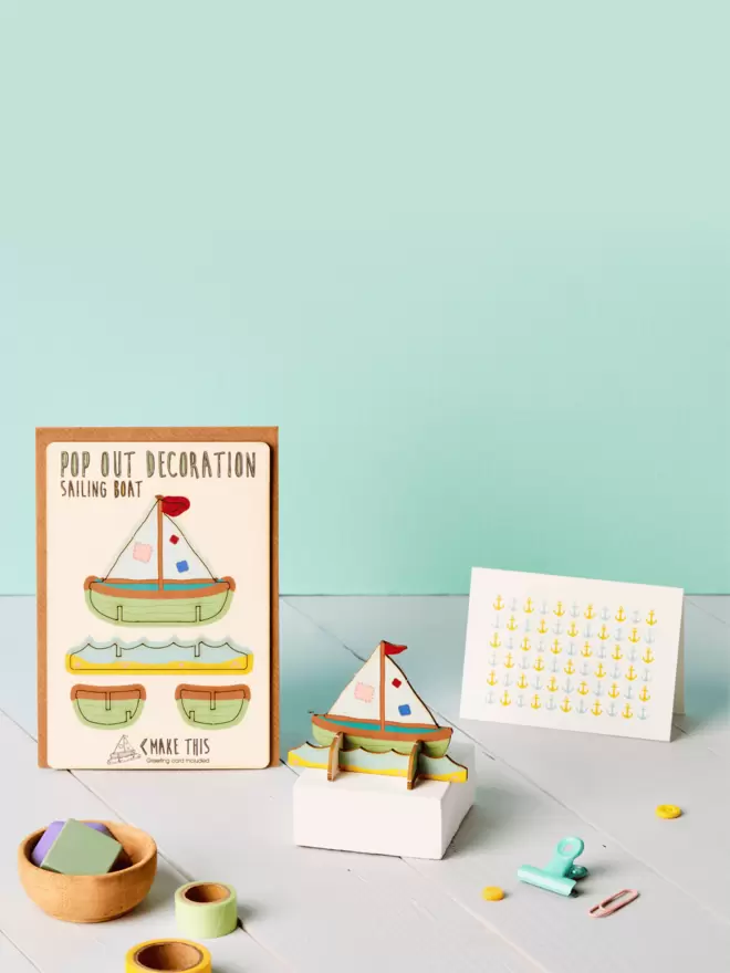 3D sailing boat decoration and anchor pattern greeting card and brown kraft envelope on top of a grey desk in front of a light green coloured background