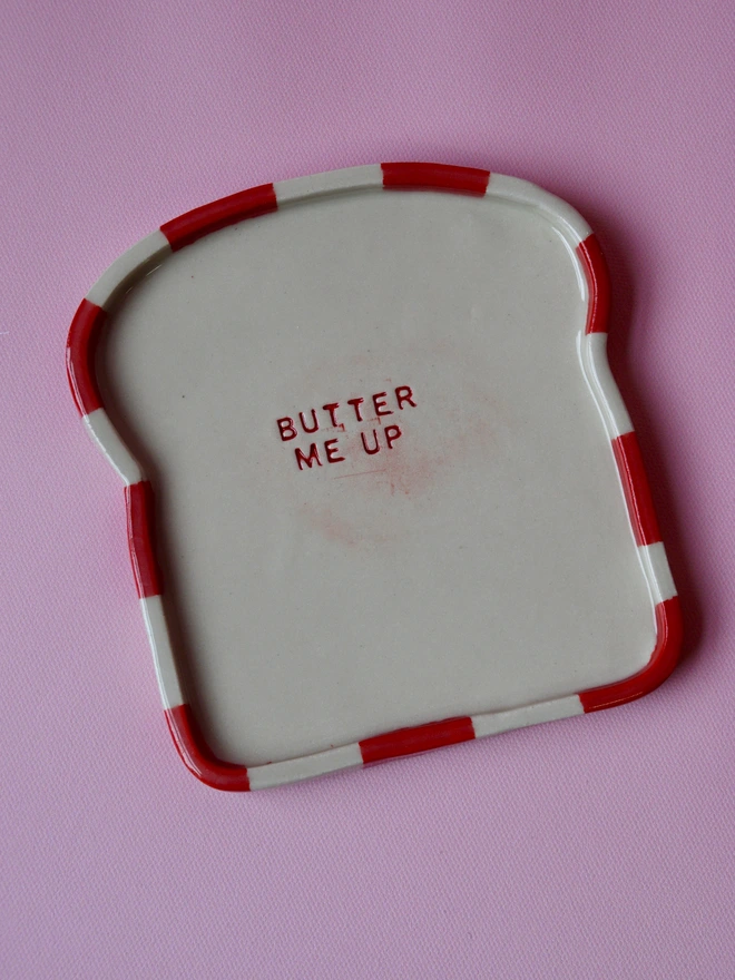 Handmade ceramic toast plate shaped like a slice of bread, featuring a red striped rim and the words “Butter Me Up” stamped in the centre, photographed on a pink background.