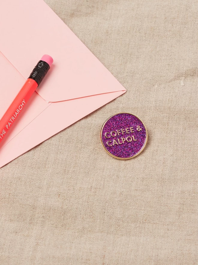 A round purple glitter enamel pin with the words Coffee and Calpol written in gold