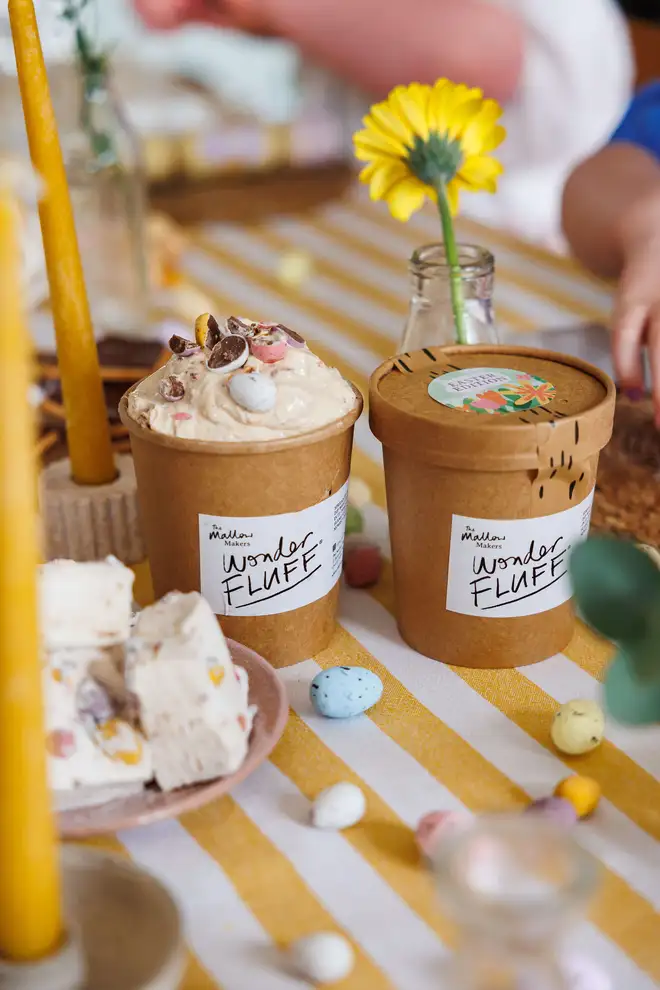Mini Eggs Wonder Fluff tubs on a striped table table cloth with a plate of mini eggs jumbo marshmallows and mini eggs scattered around