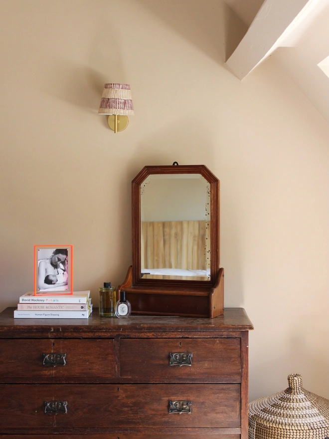 Burgundy and pink striped lampshade mounted on a brass wall fitting, bold lampshade styled above a vintage wooden chest of drawers with framed artwork and decorative accents.