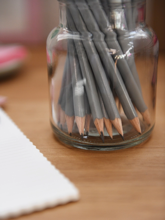 A glass jar full of grey pencils stands on a wooden desk surrounded by various stationery items.