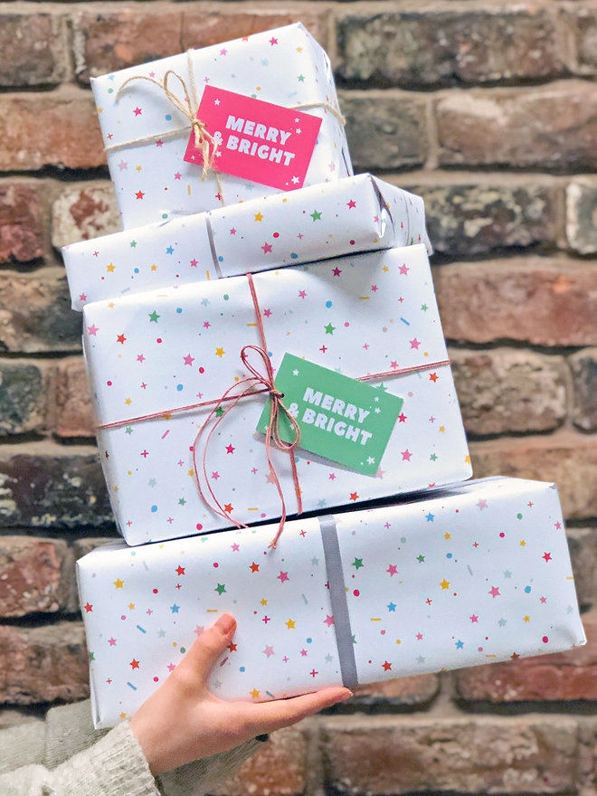 A stack of presents wrapped in pastel star wrapping paper is being held in front of a brick wall.