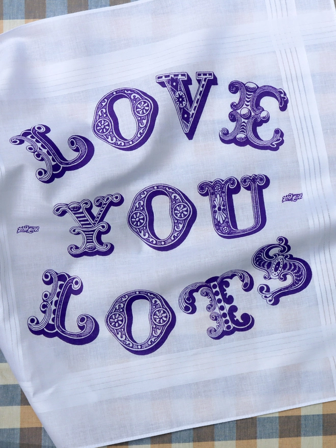 A Mr.PS Love you Lots hankie printed in violet laid flat on a gingham tablecloth