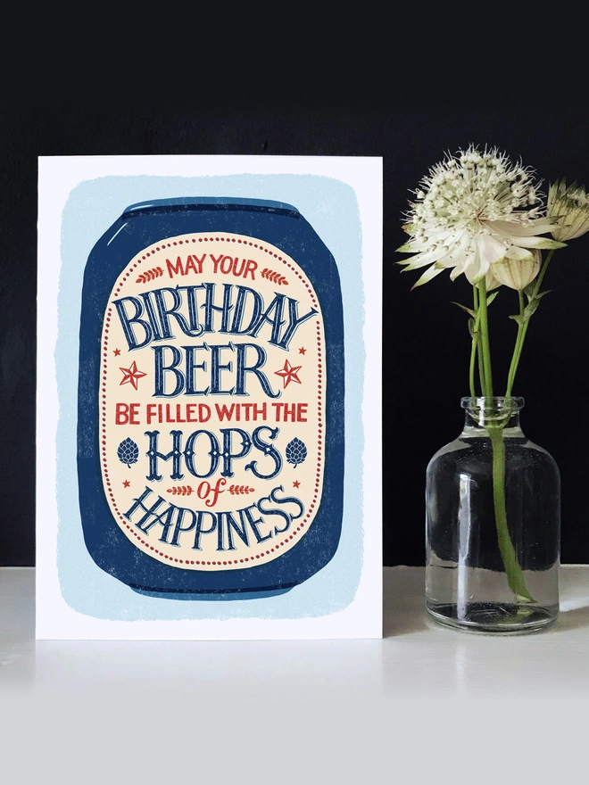 Illustrated beer can with hand lettered beer hops quote on a blue background with a white flower in a vase