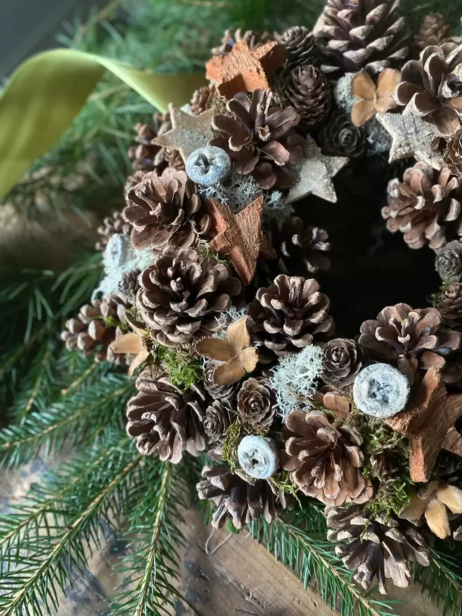 Pine Cone Wreath decorated with natural bark stars, sits on a wooden table, green foliage and a lighter thick green ribbon surrounds the wreath.