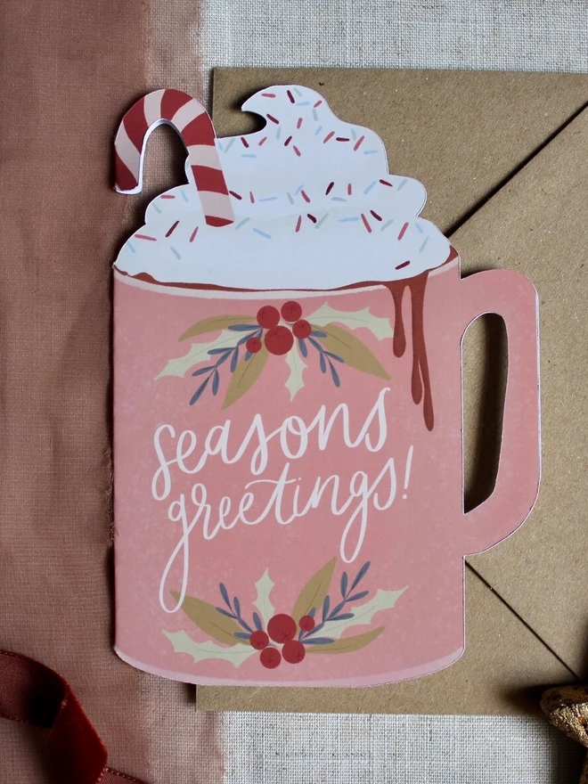 season's greetings christmas hot chocolate card. christmas card shaped like a pink mug with whipped cream on top.