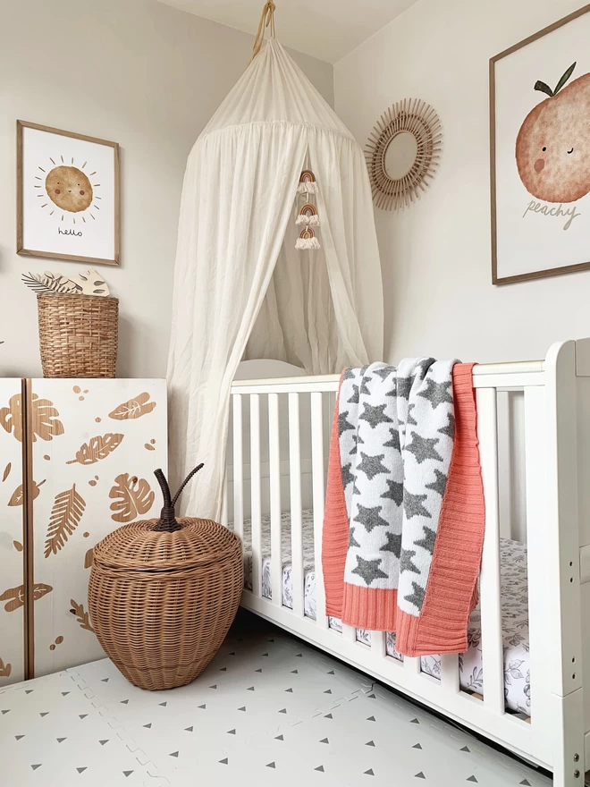 A neutral scandi style nursery with lots of natural wood, wicker accessories and a white cot. Over the cot is draped a grey and white star blanket with coral pink trim. A poster on the wall features  a poster saying ‘peachy’.