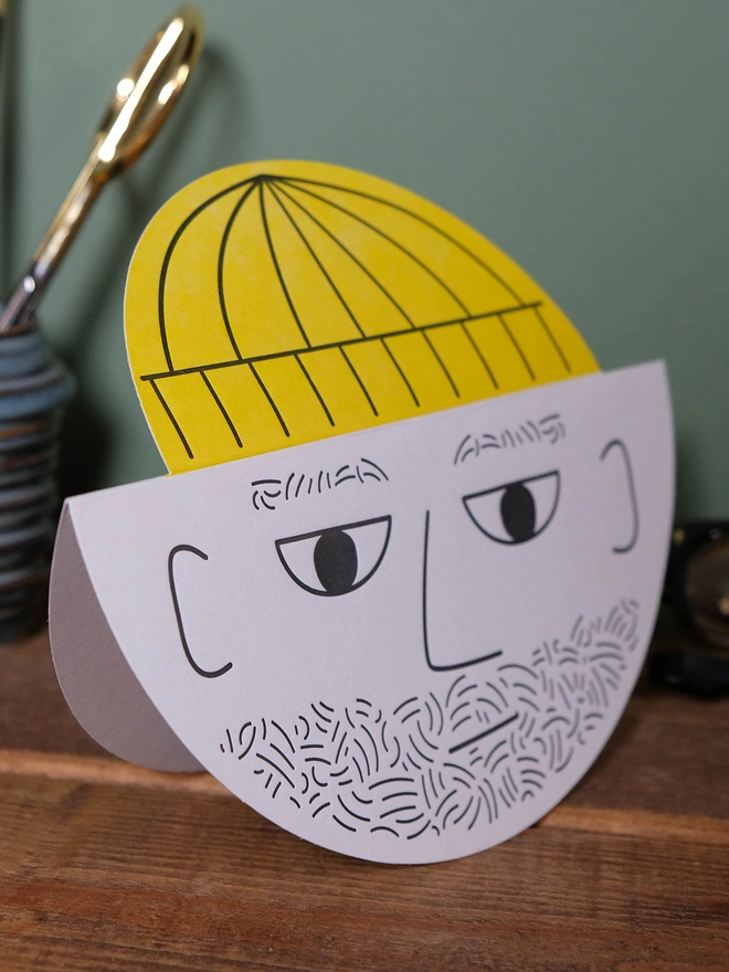 A round card with a male bearded face and yellow pop up hat. The card is hand letterpress printed in black and yellow ink on a light grey card. It is standing on a wooden shelf with green background.