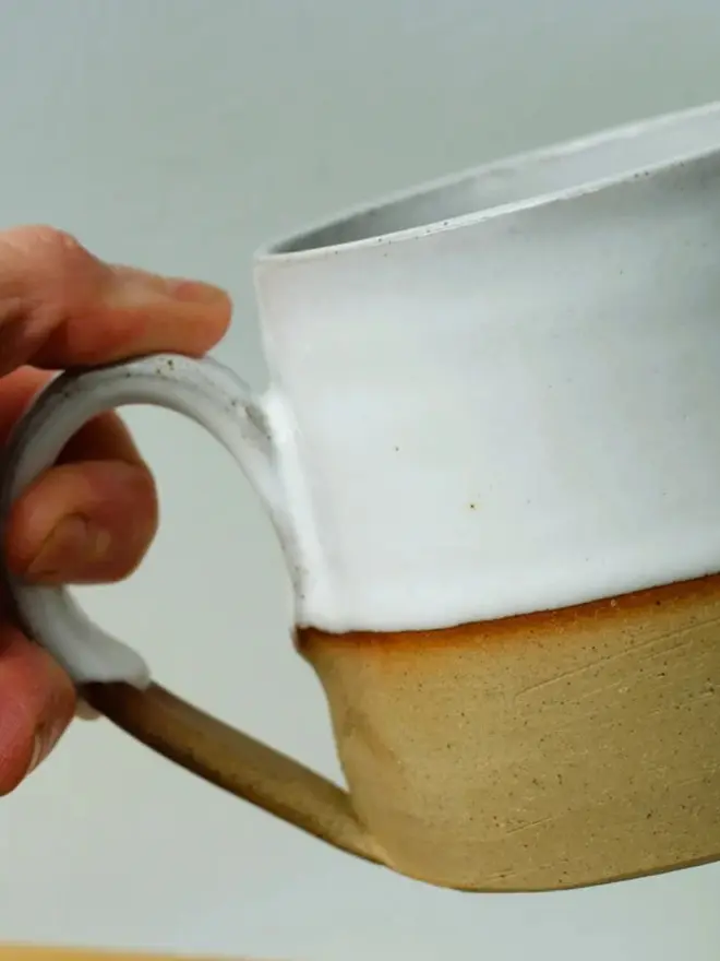 Handmade ceramic mug with a glossy white upper glaze and a natural toasted clay base, against a soft green wall background.