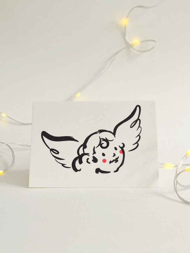 Christmas card set among fairy lights.  Card features a cute cherub character illustration in black ink with red cheeks. Cream coloured paper and background.