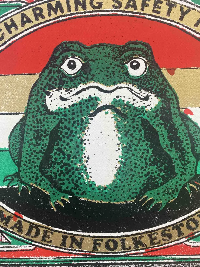  An image of a limited edition A2 screenprint. The print depicts a retro-style matchbox illustration with a central image of a green frog with a white belly, set against a background of horizontal red, white, and green stripes. The frog is framed by a gold-bordered oval. Text at the top of the oval reads "PRINCE CHARMING SAFETY MATCHES," and at the bottom, "MADE IN FOLKESTONE." The matchbox border is red with ornate details and a green trim. The match tips on the left are a textured gold. Below the main image, the print is signed and numbered 5/11. The entire image has a grainy texture, characteristic of a vintage screenprint.