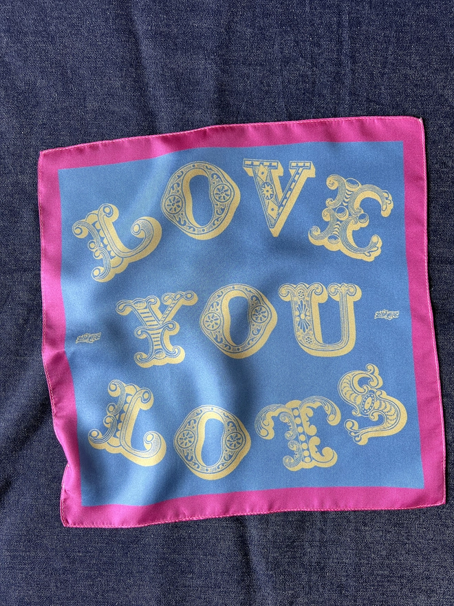 Love You Lots Silk Square blue with yellow lettering a pink border, laid on a dark cloth