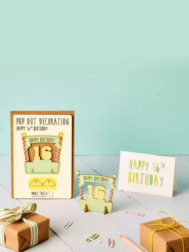 3D sixteenth birthday decoration and happy sixteenth birthday card and brown kraft envelope displayed on top of a grey desk