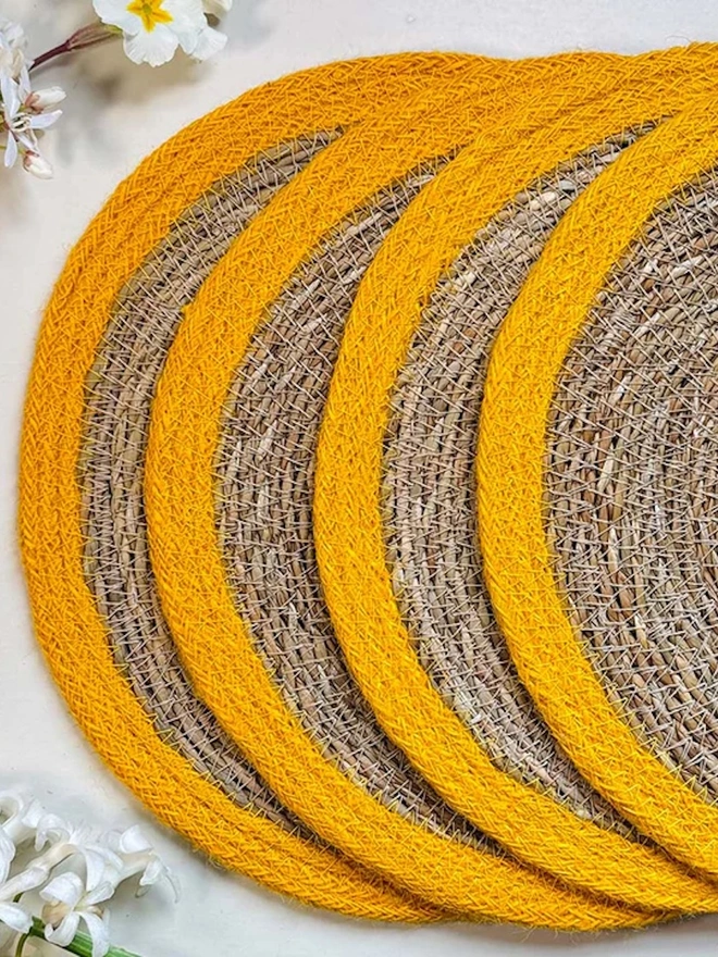 Close up of a round woven seagrass placemats with yellow rings, shown on a light surface with small white flowers.