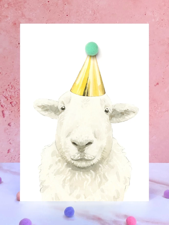 A greeting card featuring a hand painted design of a sheep, stood upright on a marble surface surrounded by pompoms. 