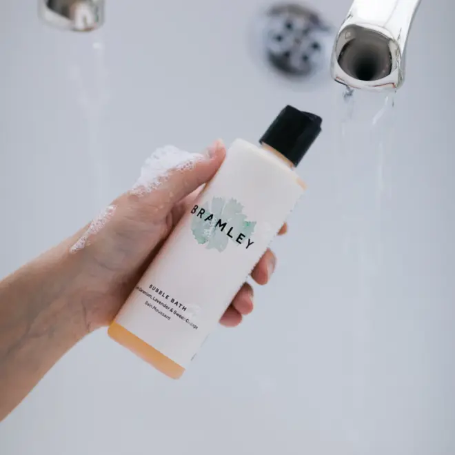 hand holding a white bottle of bubble bath with a black lid, above a white bathtub
