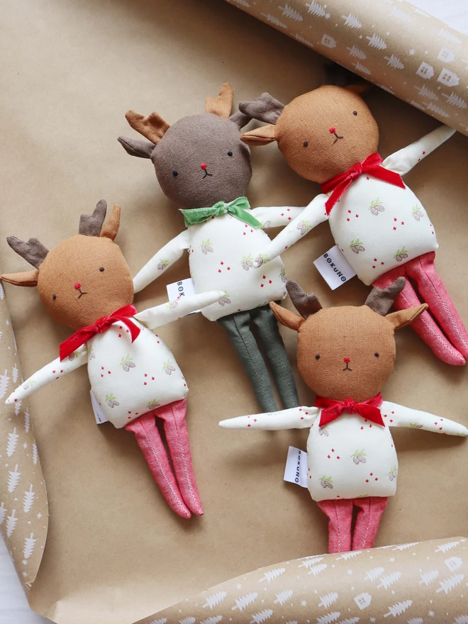Four handmade reindeer dolls arranged on brown wrapping paper with white festive tree patterns. Each doll has soft antlers, embroidered facial features, and a festive pinecone print outfit with either a red or green velvet bow. Two have green legs, and two have sparkly red legs. The dolls have "BOKUNO" fabric tags and are styled as if ready for gift wrapping.