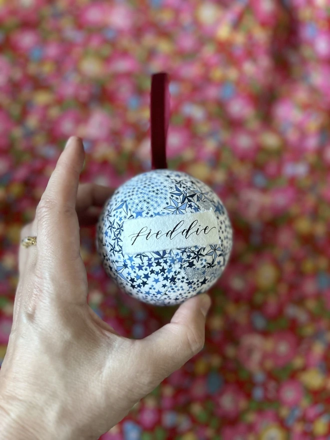 Liberty fabric blue stars Christmas bauble with Freddie handwritten on label held in hand against fabric backdrop