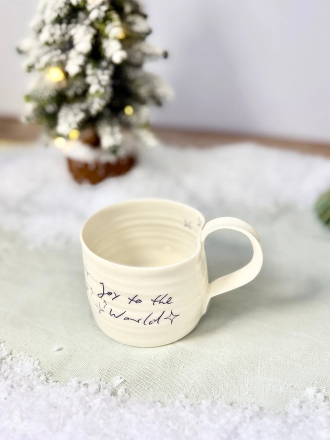 cream hand thrown porcelain mug with the words 'Joy to the World' printed onto the side with pretty line drawn stars. Sitting on a tabletop with snow, a mini lit Christmas tree 