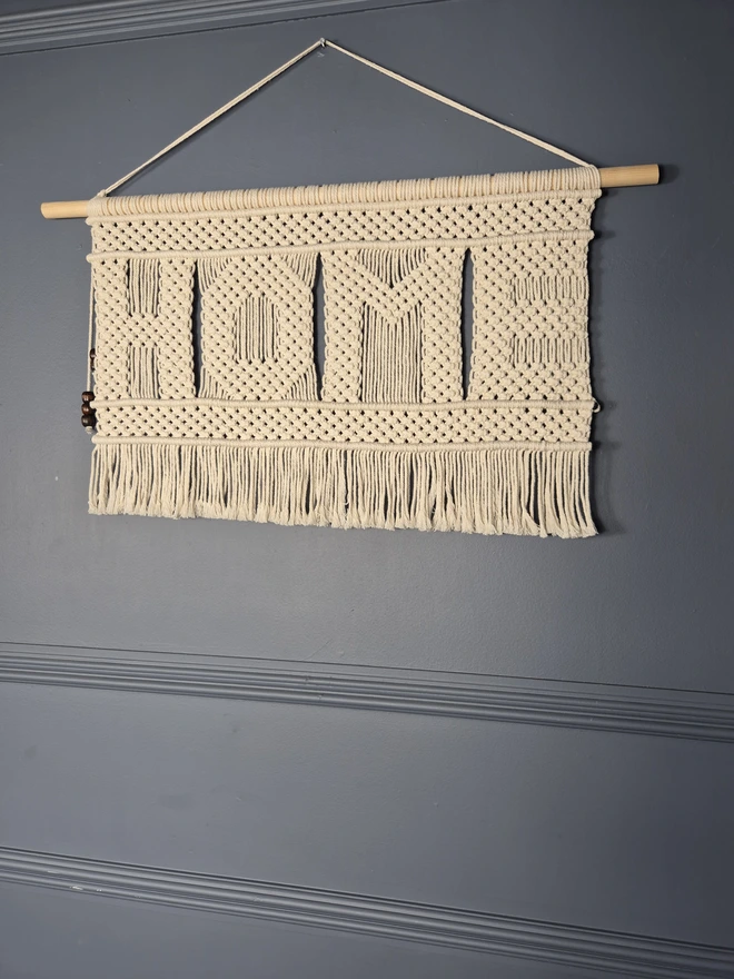 Handmade macrame wall hanger featuring the word 'HOME' intricately woven in square knots, perfect for enhancing home decor with a warm and inviting touch. A unique and one-of-a-kind wall hanger