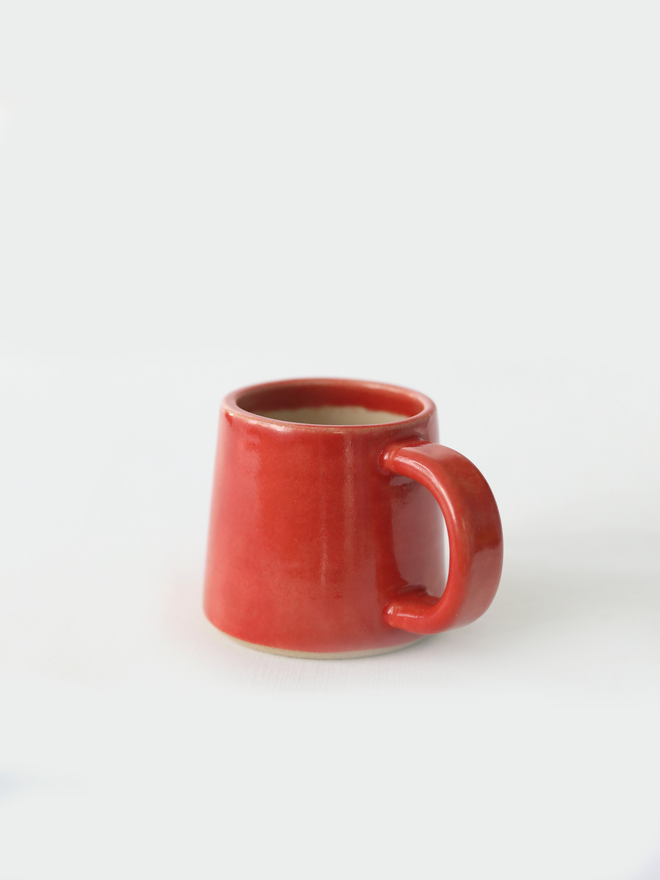 A handmade ceramic espresso mug in cherry red glaze, with a chunky loop handle, photographed against a plain light grey background