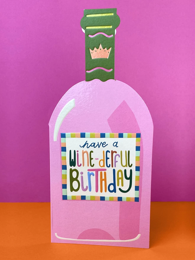 A vibrant die cut birthday card in the shape of a pink and olive wine bottle with a rainbow coloured ‘Have a wine-derful Birthday’ message