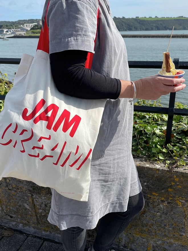 Woman in grey, stood looking out to sea holding an icecream in a pot, with a tote bag over her shoulder with JAM written above CREAM in red ink screenprinted on cream bag with red handles.