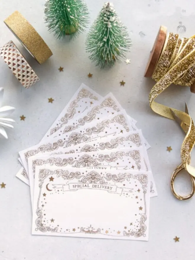 A flat lay of blank "Special Delivery" Christmas notelets with gold foil borders, surrounded by miniature frosted Christmas trees, gold glitter ribbon, washi tape, gold scissors and scattered gold star confetti.
