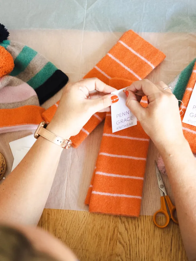 Orange wristwarmers on a table being tagged by hands and ready to be packed.