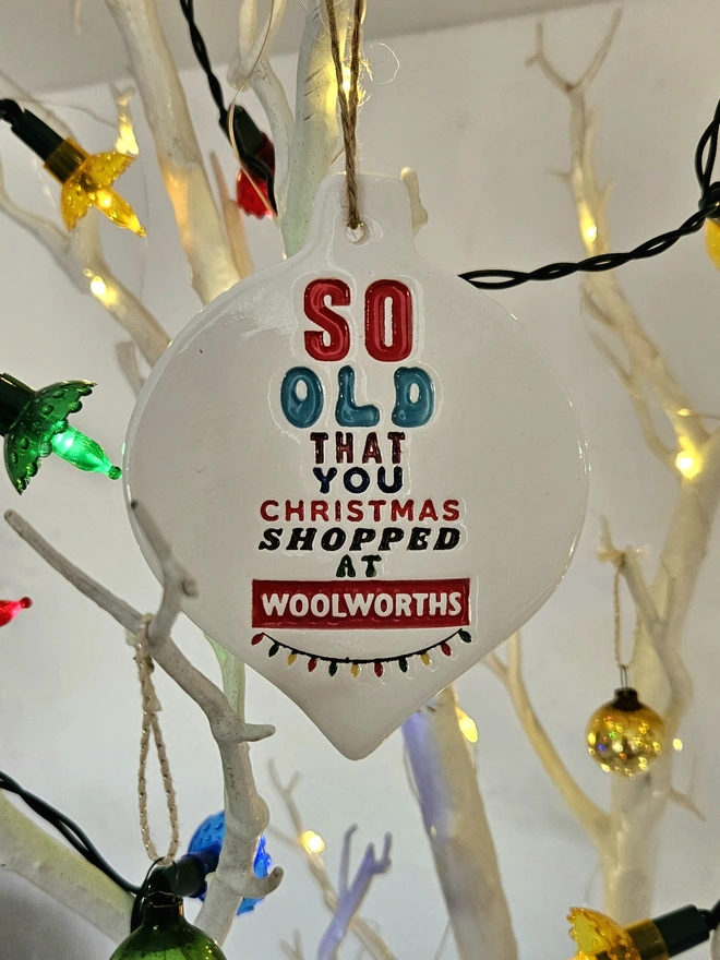 A handmade flat white ceramic bauble which measures 10cm 
 by 8cm with So old that you Christmas shopped at Woolworths written on it in colourful glazes and retro fonts with a little string of colourful fairy lights stamped into the clay.