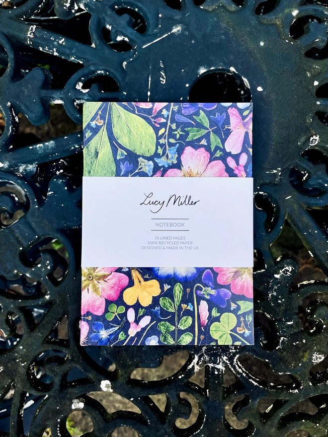 Compact Pocket-Sized Notebook with Pressed Flower Design, 'Lucy Miller' Branded Belly Band, Laying Flat on Wrought Iron Garden Table