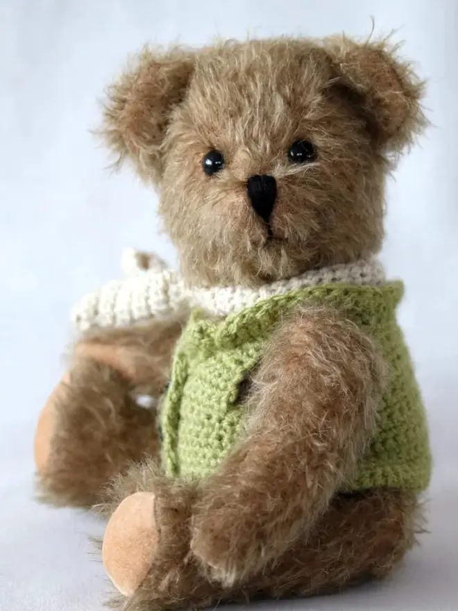 Handmade brown teddy bear with a white scarf wearing a green cardigan, sitting against a plain background.