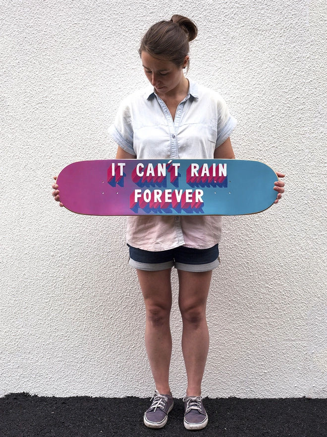 Woman holding skateboard with spray painted pink and blue background and 3d typography that reads It Can’t Rain Forever in pink and white by artist Survival Techniques. A white wall in the background and black floor.