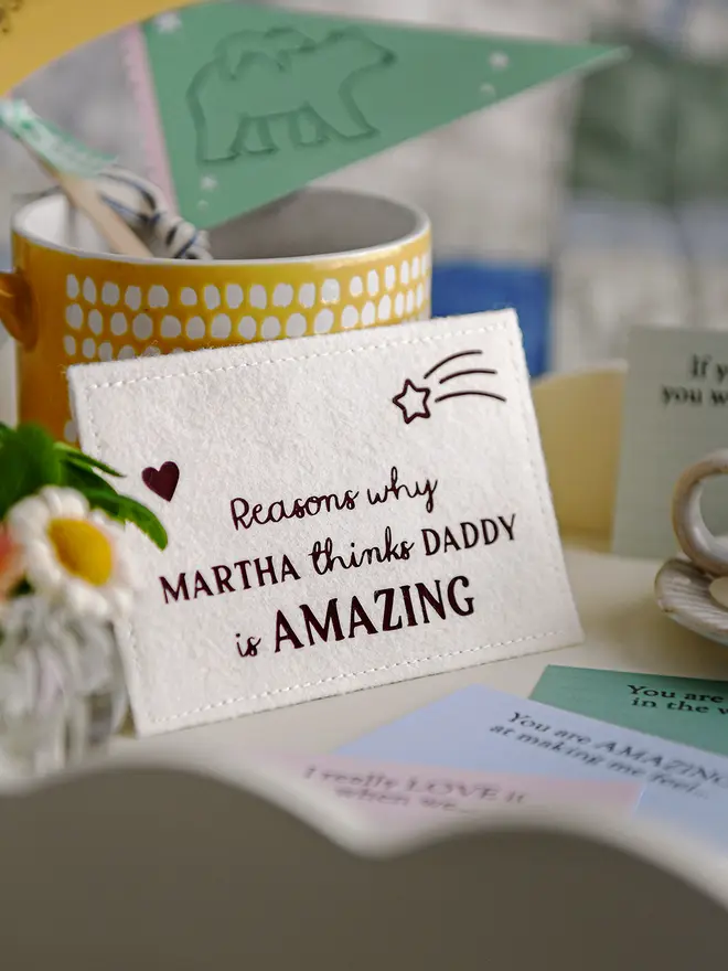 A close-up of a handmade felt envelope with the words "Reasons why MARTHA thinks DADDY is AMAZING" displayed on a white breakfast tray. Around it are pastel-coloured prompt cards with handwritten messages, a small vase of daisies, a yellow mug with crafting items, and a miniature candle on a ceramic plate, creating a cosy Father's Day setting.