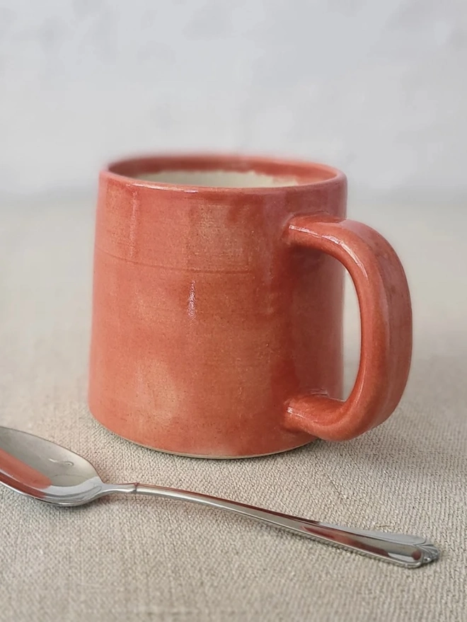 Ceramic Standard Mug, a colourful ceramic mug sitting on a plain surface against a grey backdrop. It is accompanied by a spoon. 