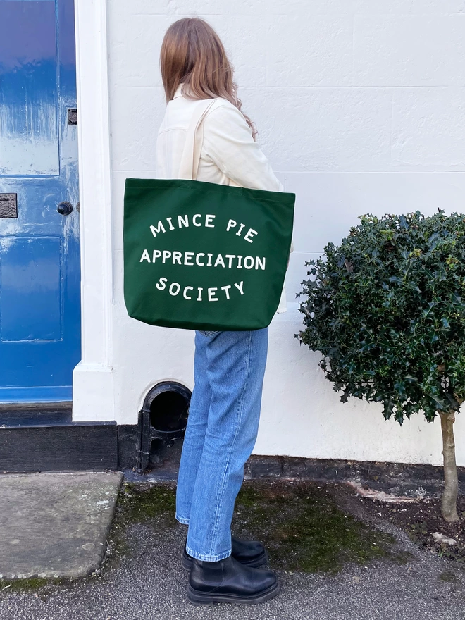 Model carrying a green canvas tote bag with 'Mince Pie Appreciation Society' printed on the front